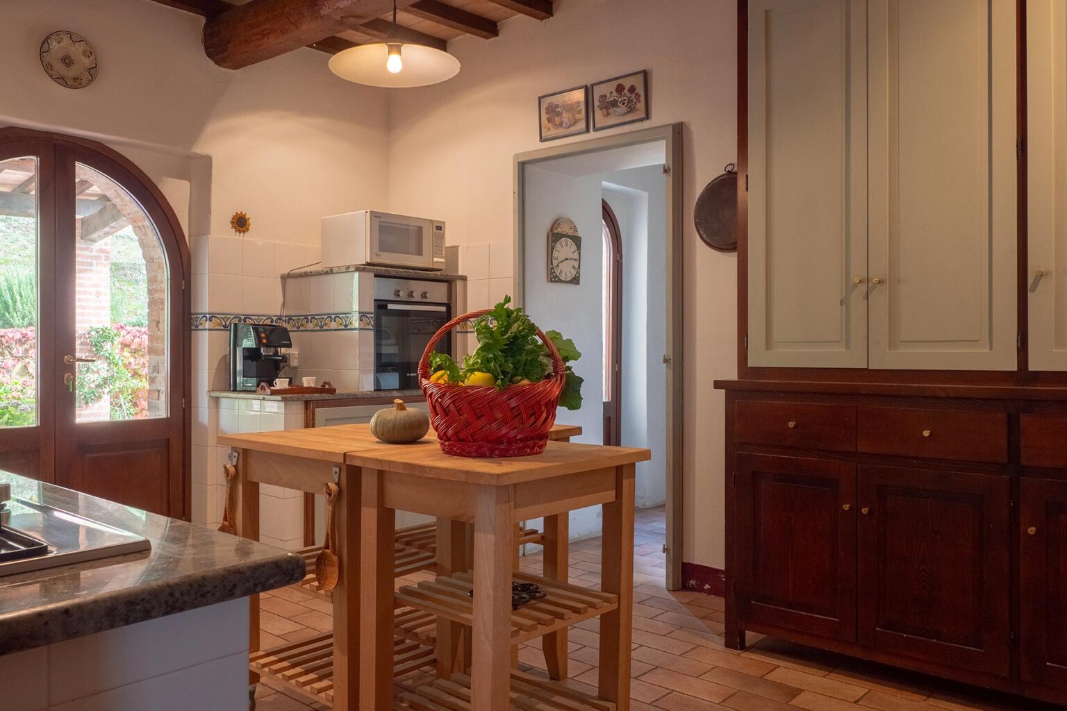 Cozy kitchen interior featuring a wooden dining table with a basket of fresh vegetables, modern appliances, and traditional cabinetry, illuminated by natural light from a window. | Only on SopranoVillas