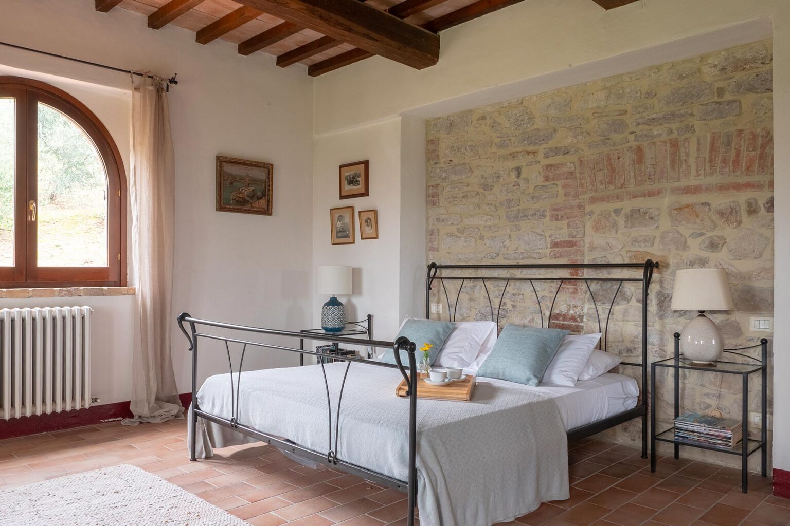 Cozy bedroom featuring a wrought iron bed with soft bedding, a rustic stone wall, and natural light from a large arched window, creating a warm and inviting atmosphere. | Only on SopranoVillas