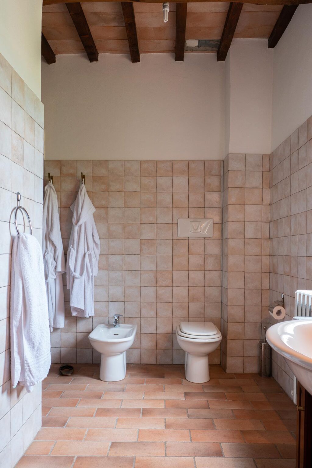 Modern bathroom featuring beige tiled walls, two hanging white bathrobes, and essential fixtures including a toilet and a towel rack. Ideal for showcasing home decor and design ideas. | Only on SopranoVillas