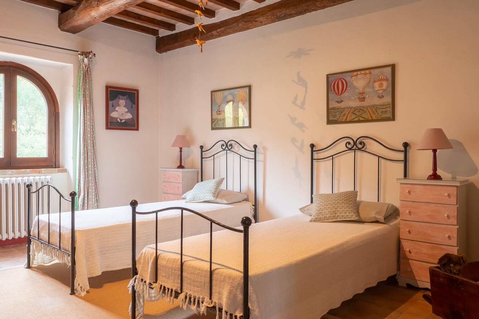 Cozy bedroom featuring two metal frame twin beds, pastel pink nightstands, and rustic wooden beams, adorned with framed artwork and soft natural light from a large window. | Only on SopranoVillas