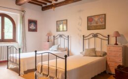 Cozy bedroom featuring two metal frame twin beds, pastel pink nightstands, and rustic wooden beams, adorned with framed artwork and soft natural light from a large window.