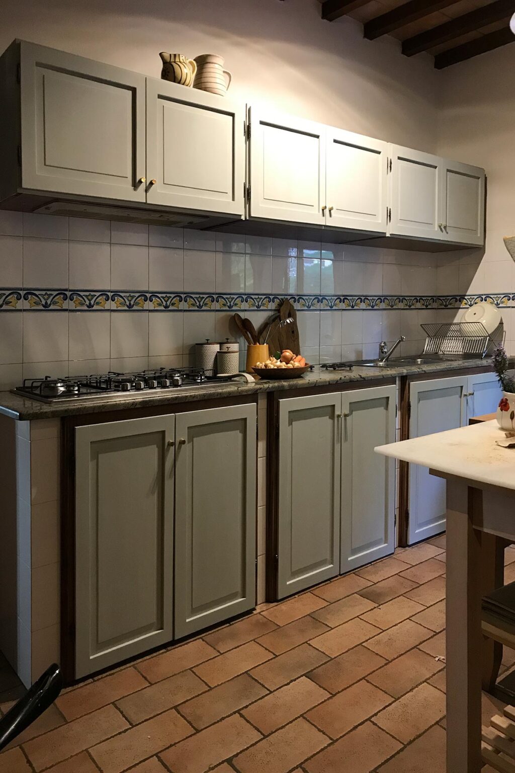 Modern kitchen interior featuring a sleek granite countertop, gas stove, and wooden cabinets with a light green finish. The tiled backsplash adds a decorative touch, while kitchen utensils and fresh fruit create an inviting atmosphere. | Only on SopranoVillas