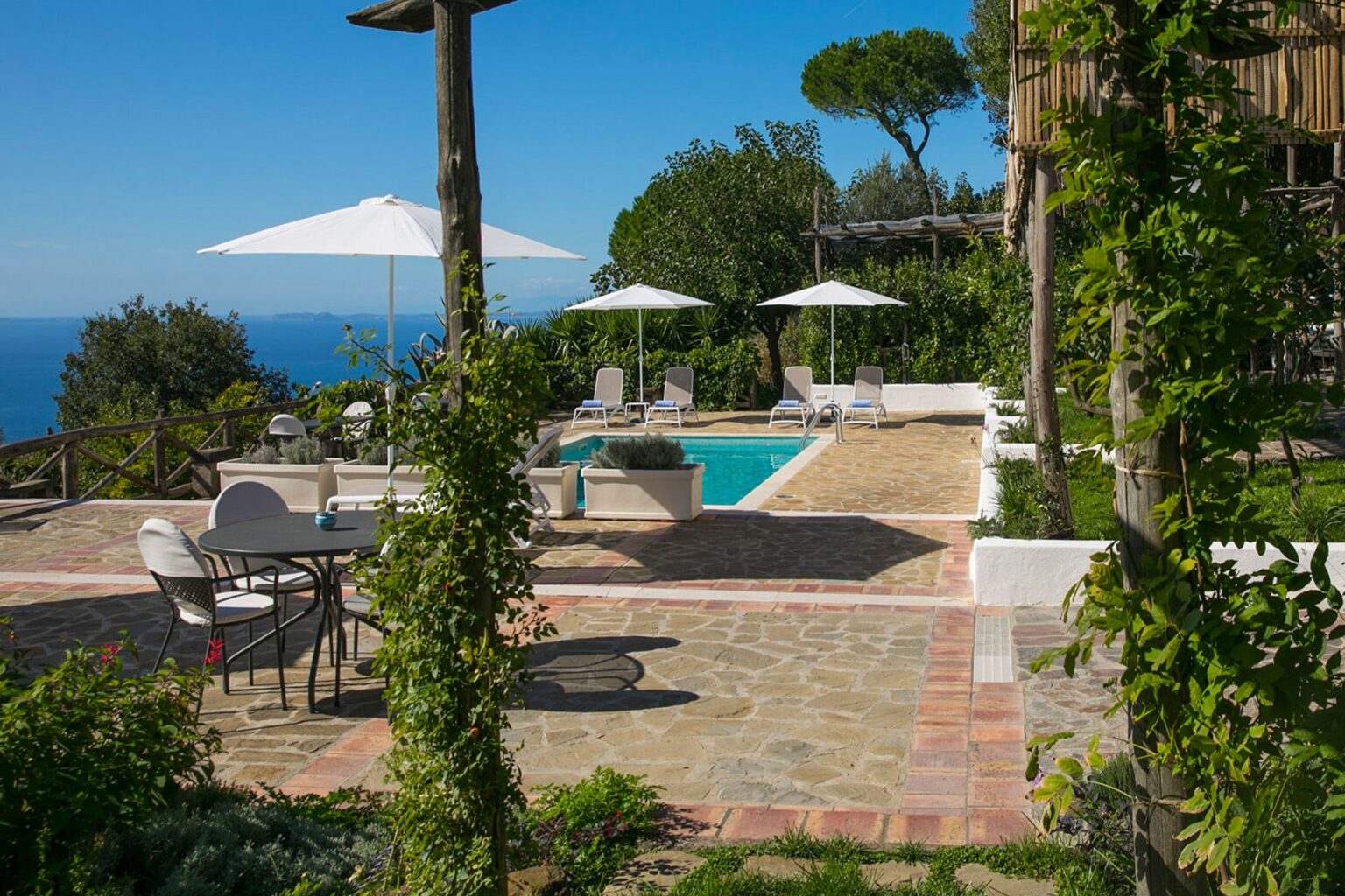 Luxurious outdoor pool area in a villa with sun loungers and umbrellas, surrounded by lush greenery and scenic ocean views. Ideal for relaxation and leisure in a Mediterranean setting. | Only on SopranoVillas