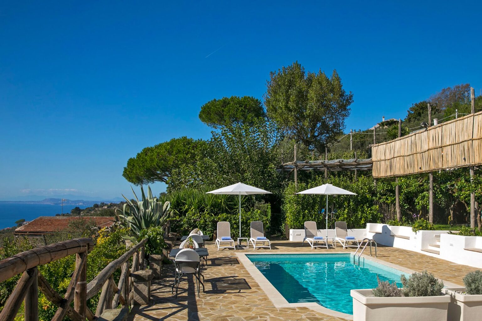 Luxurious outdoor pool area with sun loungers and parasols, surrounded by lush greenery and a wooden deck, under a clear blue sky. Ideal for relaxation and enjoying scenic views. | Only on SopranoVillas