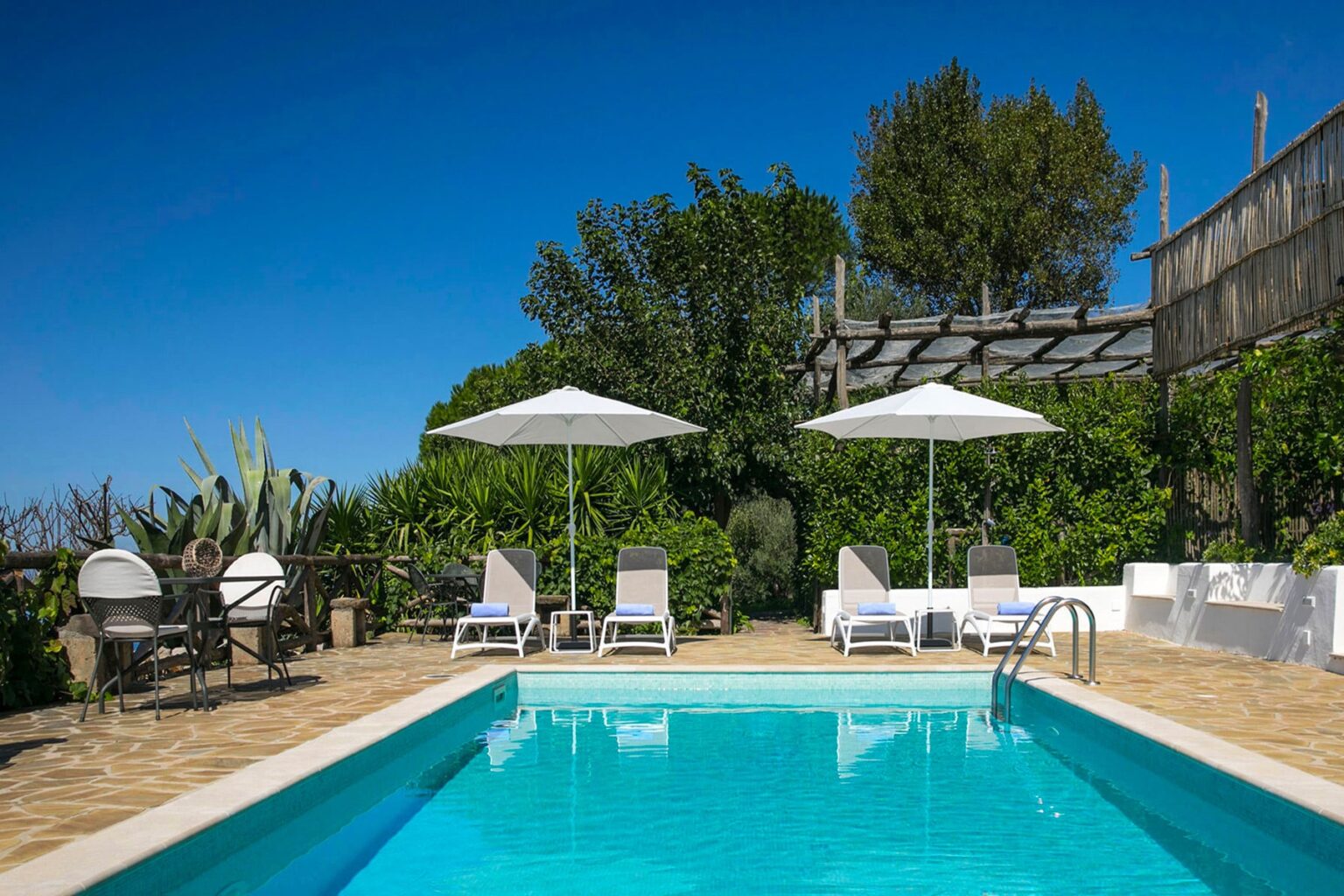 Serene outdoor swimming pool surrounded by lush greenery, featuring sun loungers and umbrellas under a clear blue sky, ideal for relaxation and leisure. | Only on SopranoVillas