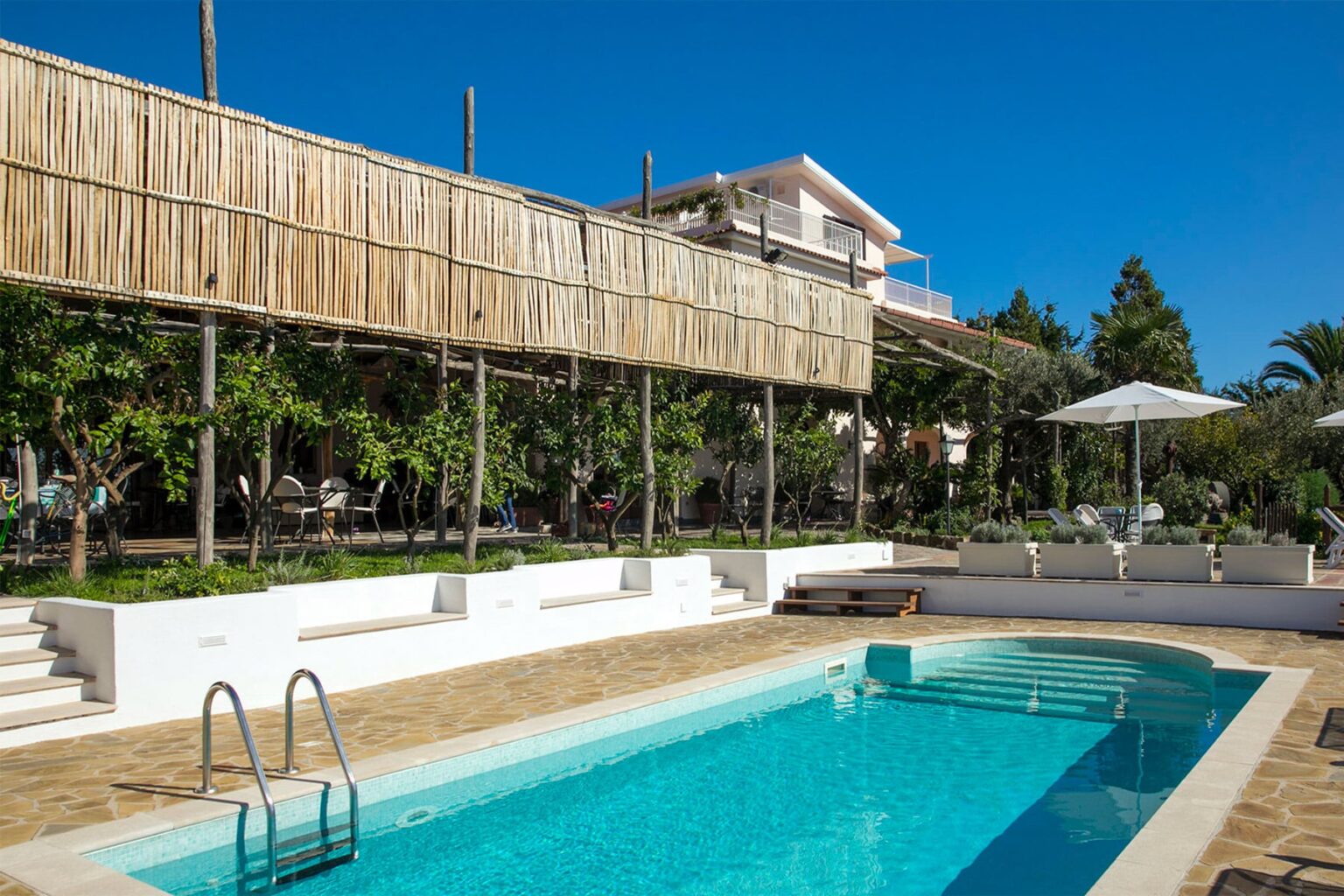 Modern outdoor swimming pool with a wooden deck, surrounded by lush greenery and a sunny blue sky, featuring a white parasol and a contemporary building in the background. | Only on SopranoVillas