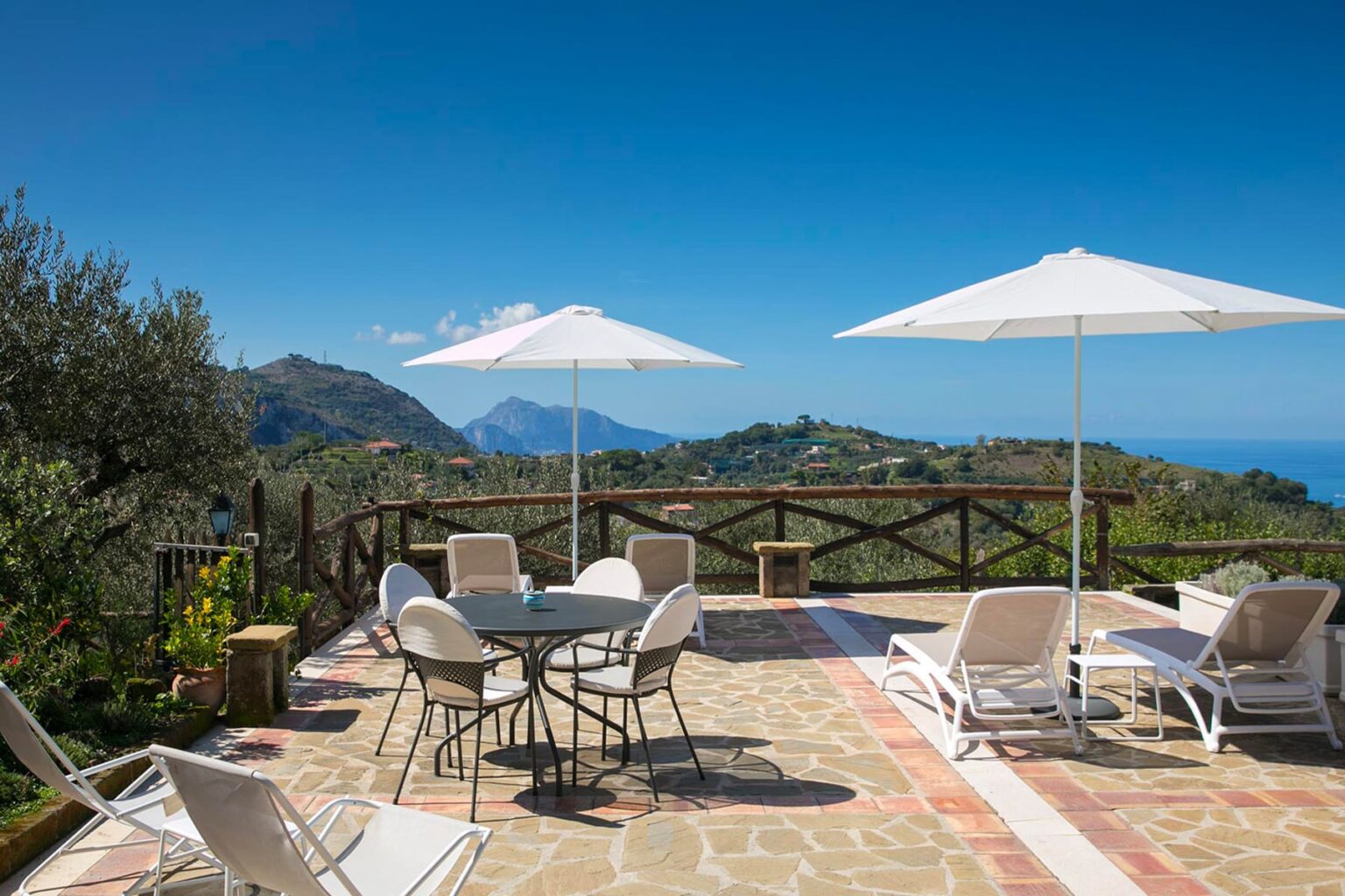 Scenic outdoor terrace with loungers and a dining table, surrounded by lush greenery and mountains, overlooking the ocean under a clear blue sky. | Only on SopranoVillas