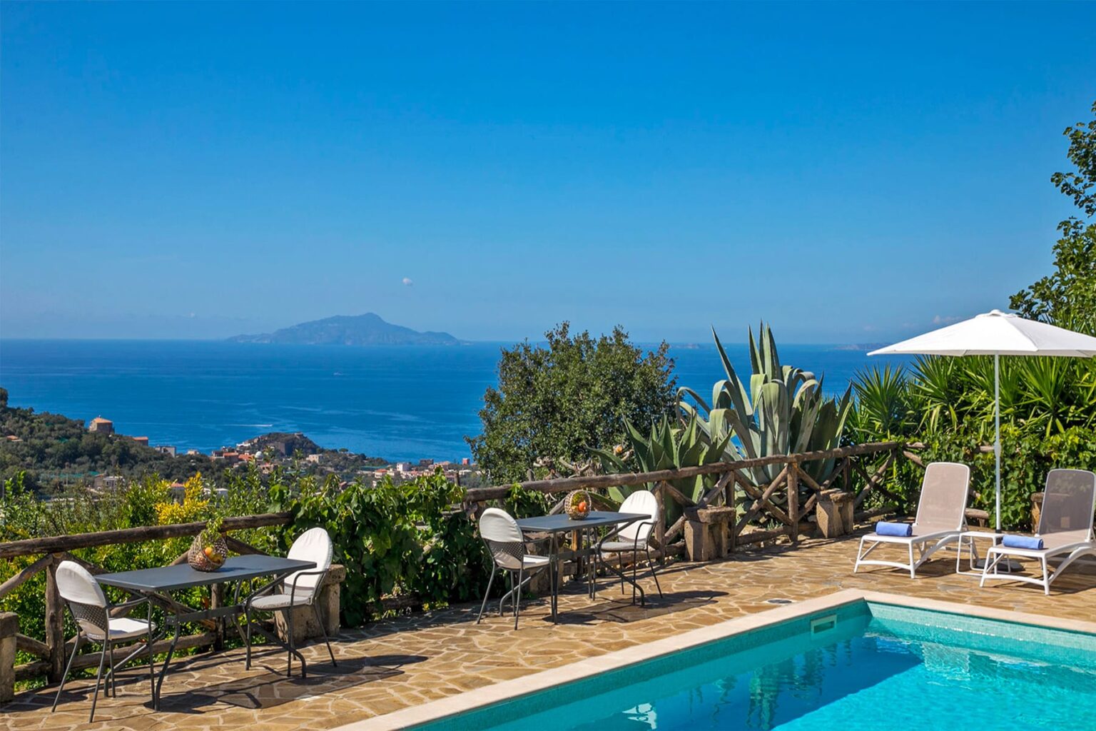 Scenic view of a tranquil poolside area with lounge chairs and a table, overlooking a vibrant blue ocean and distant islands under a clear sky. Ideal for relaxation and vacation getaways. | Only on SopranoVillas
