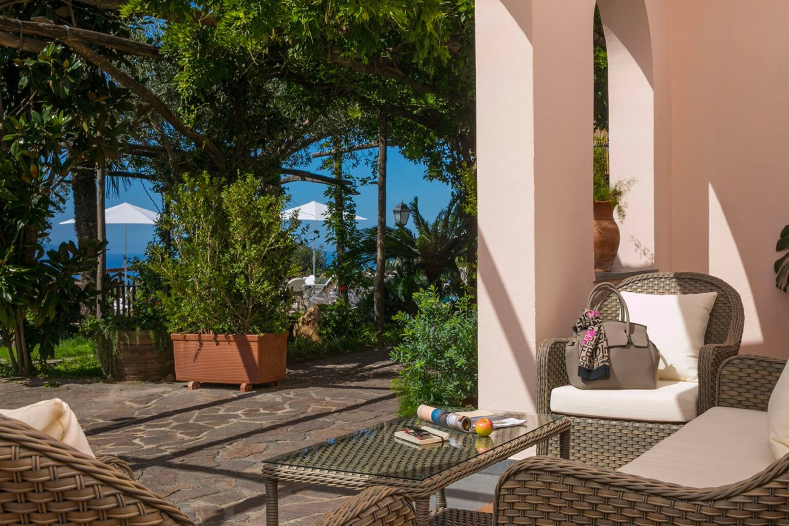 Cozy outdoor seating area with woven furniture, surrounded by lush greenery and potted plants, featuring a table with refreshments and a view of the blue sky and distant umbrellas. Perfect for relaxation and enjoying nature. | Only on SopranoVillas