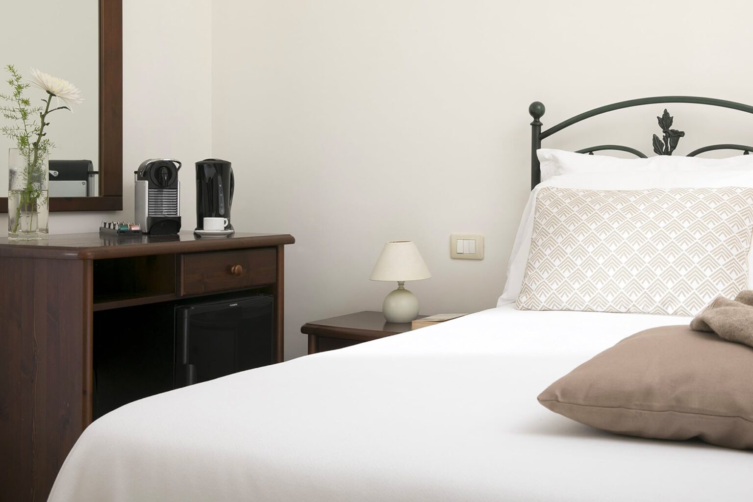 Cozy hotel room featuring a neatly made bed with a decorative pillow, a wooden nightstand with a lamp, and a coffee maker, creating a welcoming atmosphere for guests. | Only on SopranoVillas
