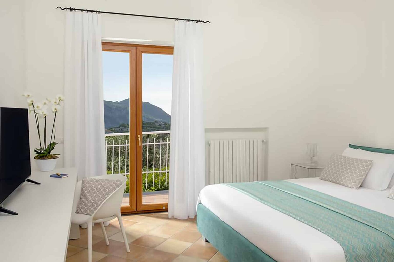 Modern hotel bedroom with a queen-sized bed, white linens, and a turquoise throw, featuring a large window with mountain views, a flat-screen TV, and minimalist decor. | Only on SopranoVillas