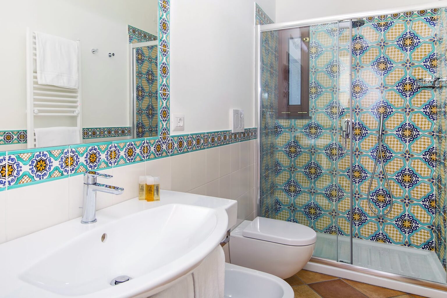 Modern bathroom featuring a sleek white sink, toilet, and a glass shower enclosure. The walls are adorned with colorful patterned tiles, complemented by a decorative border. Natural light enhances the spacious feel, creating a bright and inviting atmosphere. | Only on SopranoVillas