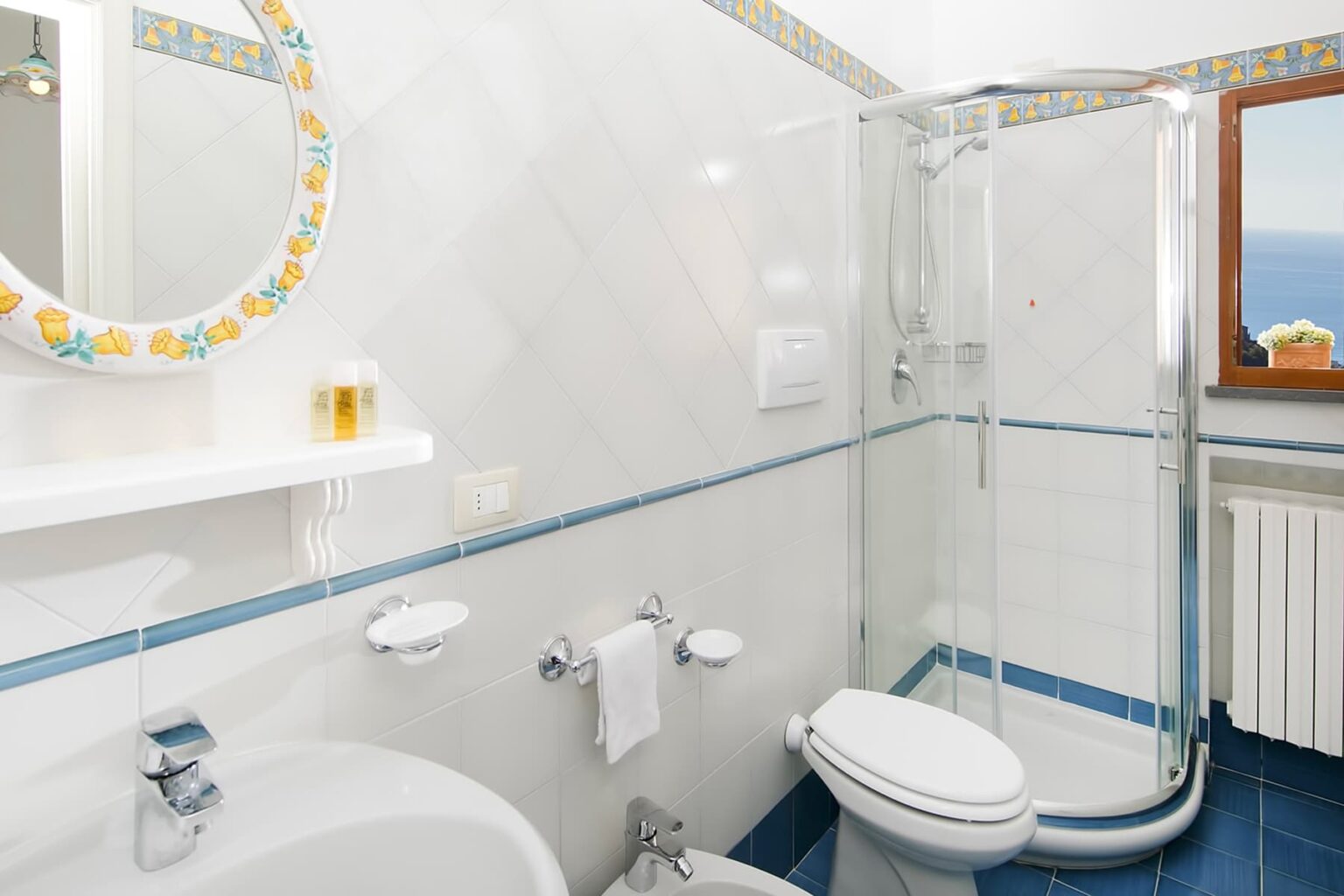 Modern bathroom featuring a glass shower, white toilet, and a sink with a mirror. The walls are tiled with a light pattern, and there are decorative elements in blue and floral designs. Natural light enters through a window, enhancing the space's brightness. | Only on SopranoVillas