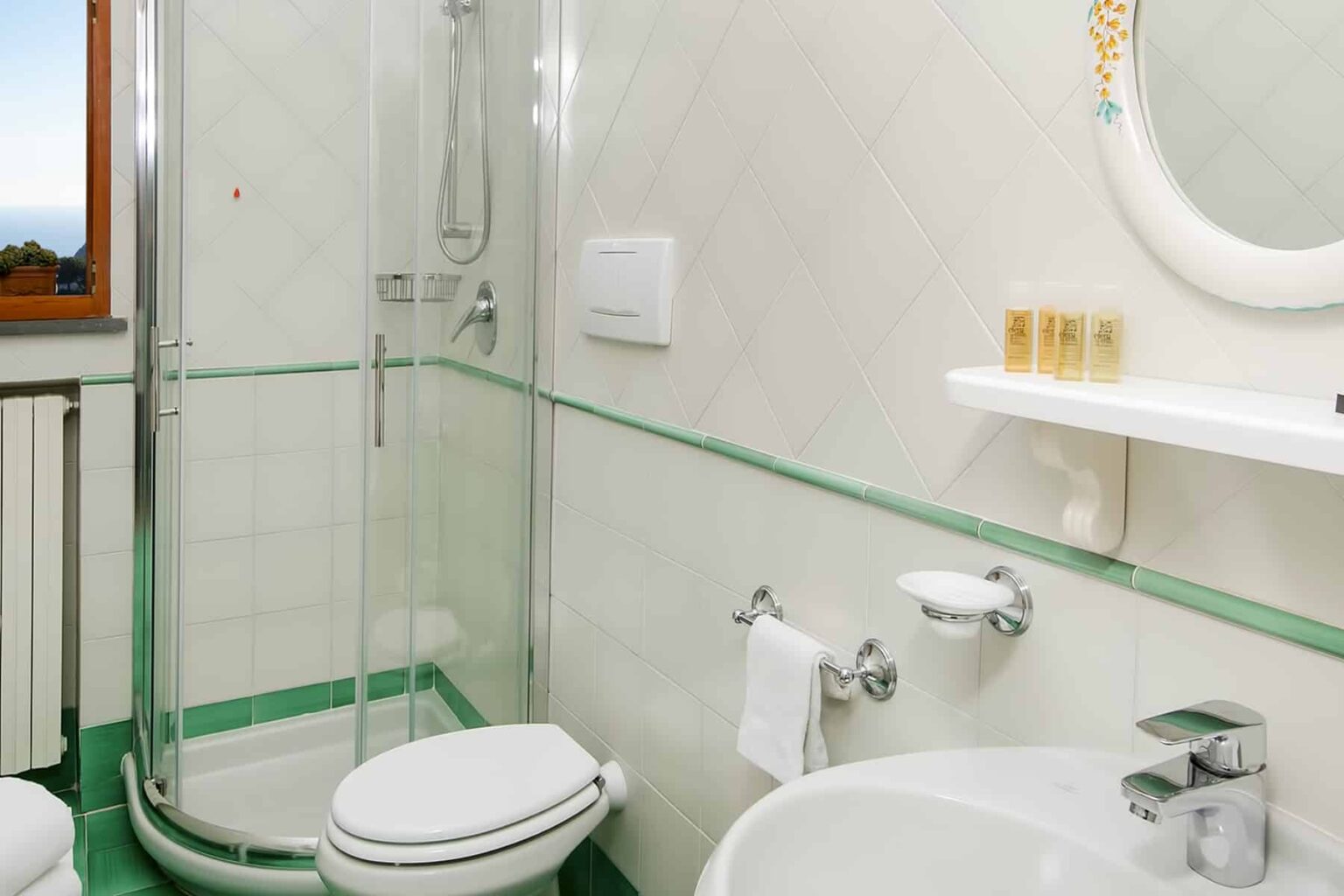 Modern bathroom featuring a glass shower enclosure, white toilet, and sink with a mirror above. The decor includes light-colored tiled walls with green accents and a shelf with toiletries. Natural light enters through a window, enhancing the space's brightness. | Only on SopranoVillas