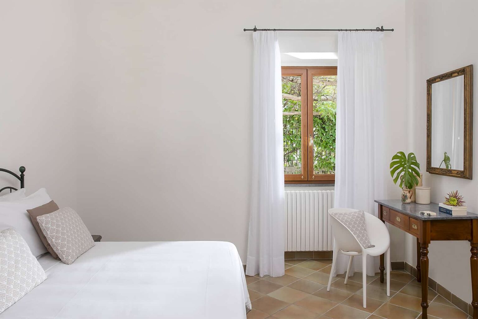 Bright and airy bedroom featuring a large window with sheer white curtains, a cozy bed with decorative pillows, a wooden vanity with a mirror, and a potted plant, creating a serene and inviting atmosphere. | Only on SopranoVillas
