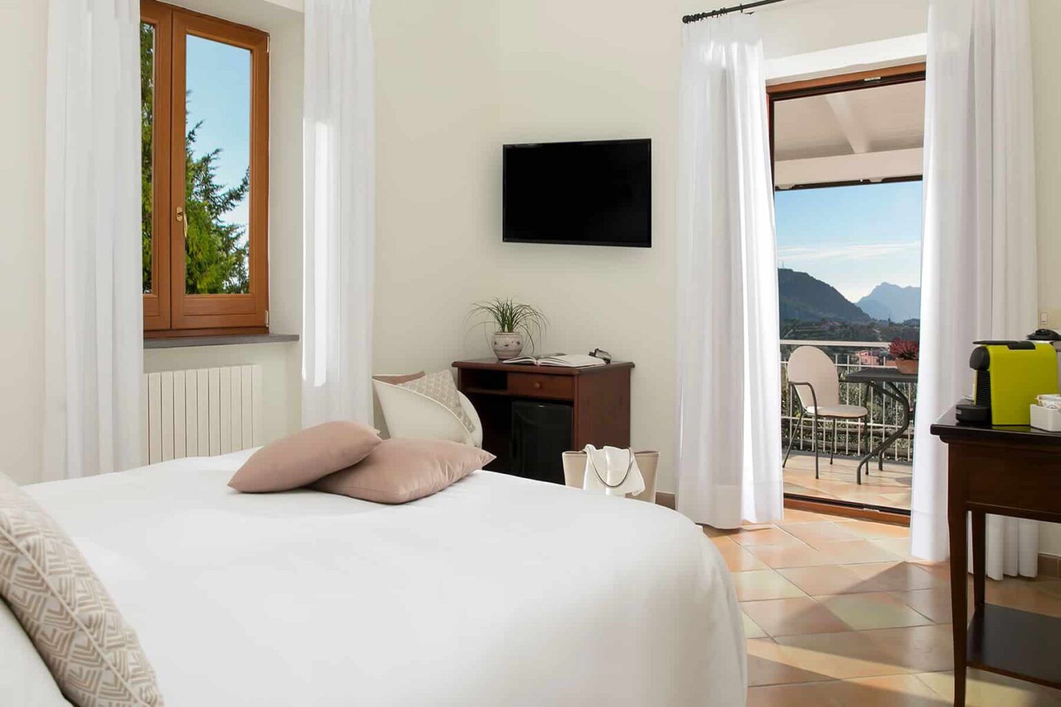 Cozy hotel room featuring a comfortable bed with decorative pillows, a flat-screen TV, and large windows offering a scenic mountain view. The room includes a small seating area and access to a balcony with outdoor seating. Ideal for relaxation and enjoying nature. | Only on SopranoVillas