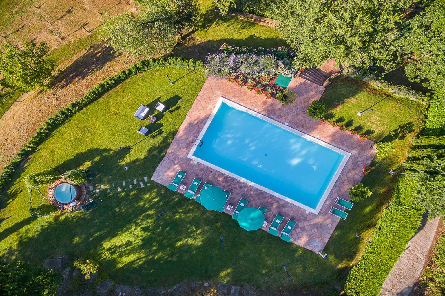 Aerial view of a rectangular swimming pool surrounded by lush green grass, lounge chairs, and a small hot tub, set in a tranquil outdoor landscape. | Only on SopranoVillas