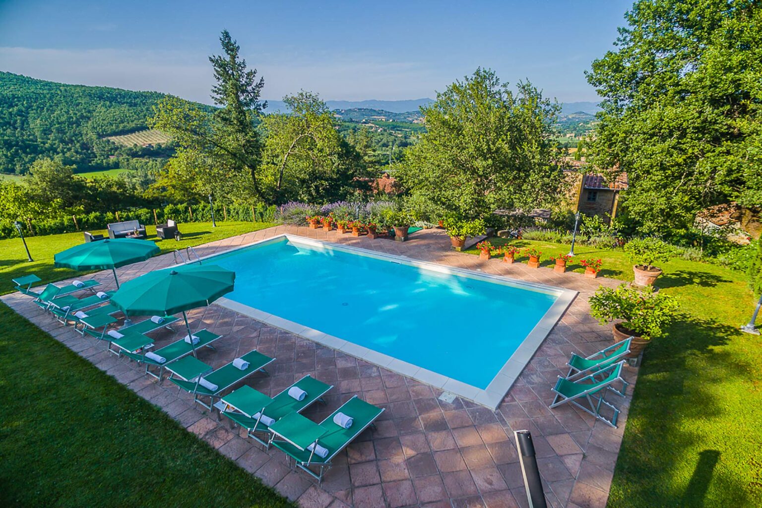 Aerial view of a serene outdoor swimming pool surrounded by lush greenery, lounge chairs, and umbrellas, set against a picturesque landscape of rolling hills and trees. Ideal for relaxation and leisure activities. | Only on SopranoVillas