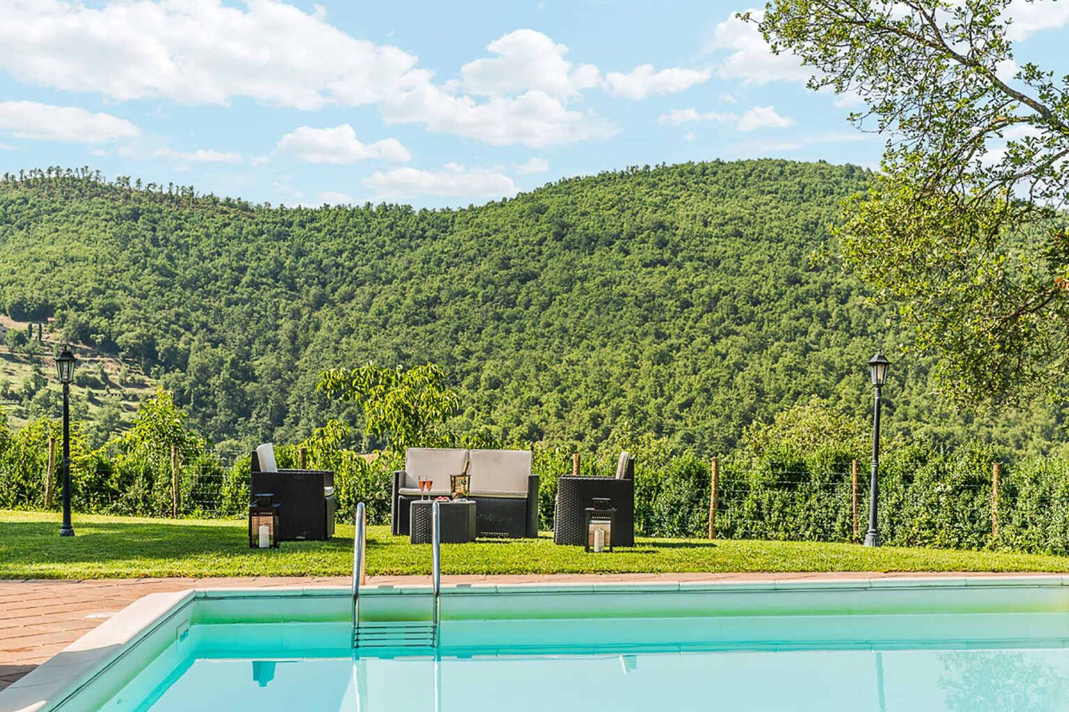 Scenic outdoor pool with lounge chairs overlooking lush green hills under a blue sky with fluffy clouds. Perfect for relaxation and enjoying nature. | Only on SopranoVillas