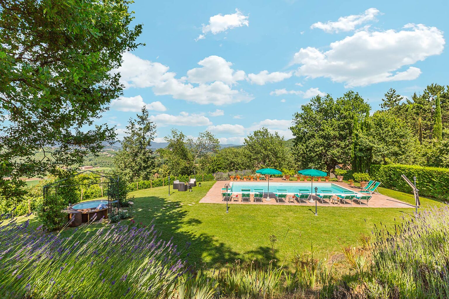 Scenic view of a tranquil outdoor pool surrounded by lush greenery and trees, with lounge chairs and umbrellas, set against a bright blue sky with fluffy clouds. Ideal for relaxation and leisure in a serene environment. | Only on SopranoVillas