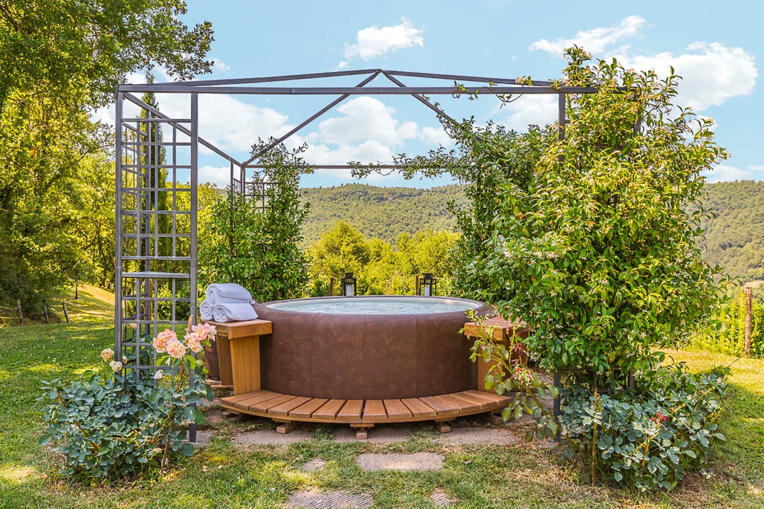 Outdoor hot tub surrounded by lush greenery and a wooden deck, set against a scenic hillside backdrop with blue skies and fluffy clouds. | Only on SopranoVillas