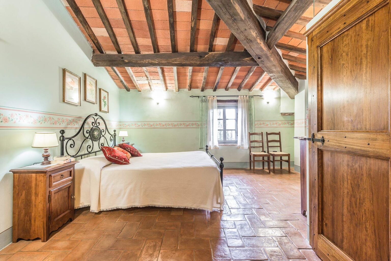 Cozy bedroom with a wooden ceiling, featuring a double bed with decorative pillows, a wooden nightstand, and two chairs. The room has a warm color palette and natural light coming from a window, creating an inviting atmosphere. | Only on SopranoVillas
