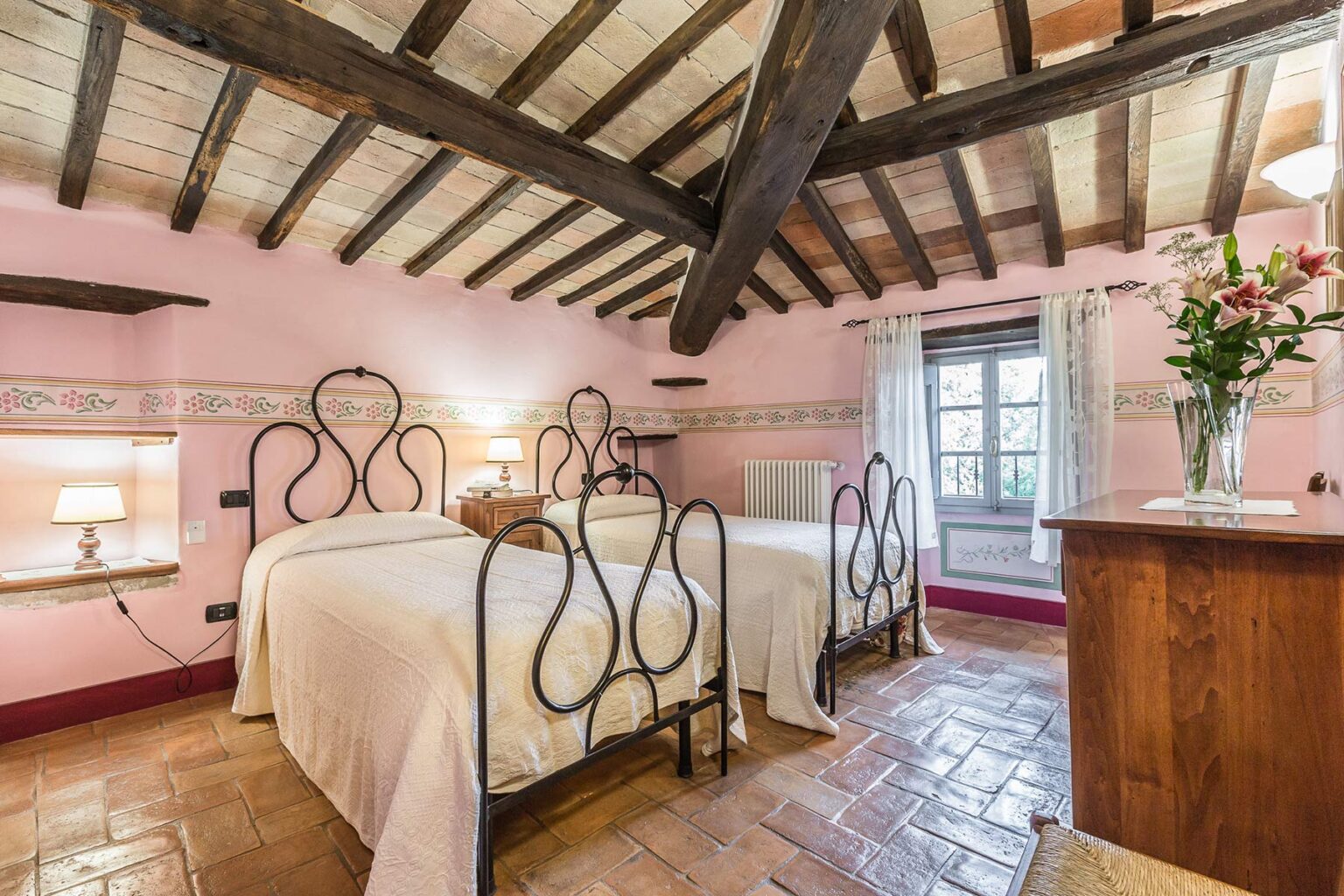 Cozy guest room featuring two wrought iron beds, pink walls, and wooden beams, with a window overlooking a scenic view and a wooden dresser with fresh flowers. Ideal for a relaxing stay. | Only on SopranoVillas