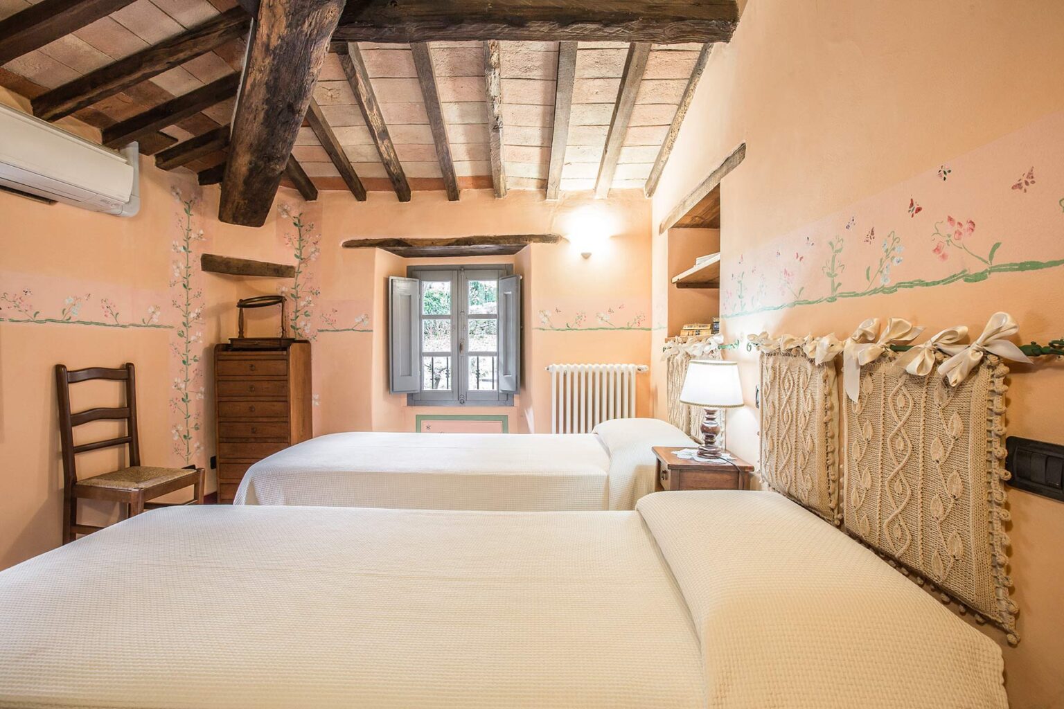 Cozy guest bedroom with two twin beds, rustic wooden beams, and warm peach-colored walls adorned with floral accents, featuring a window that invites natural light. Ideal for a relaxing stay or vacation retreat. | Only on SopranoVillas