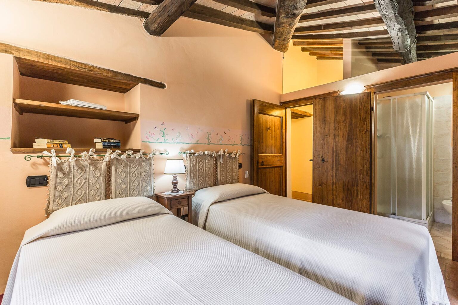 Cozy guest room featuring two twin beds with white linens, wooden shelves, and rustic decor. The room has a warm peach wall color and exposed wooden beams. A private bathroom is visible in the background, enhancing the inviting atmosphere. Perfect for travelers seeking comfort and charm. | Only on SopranoVillas