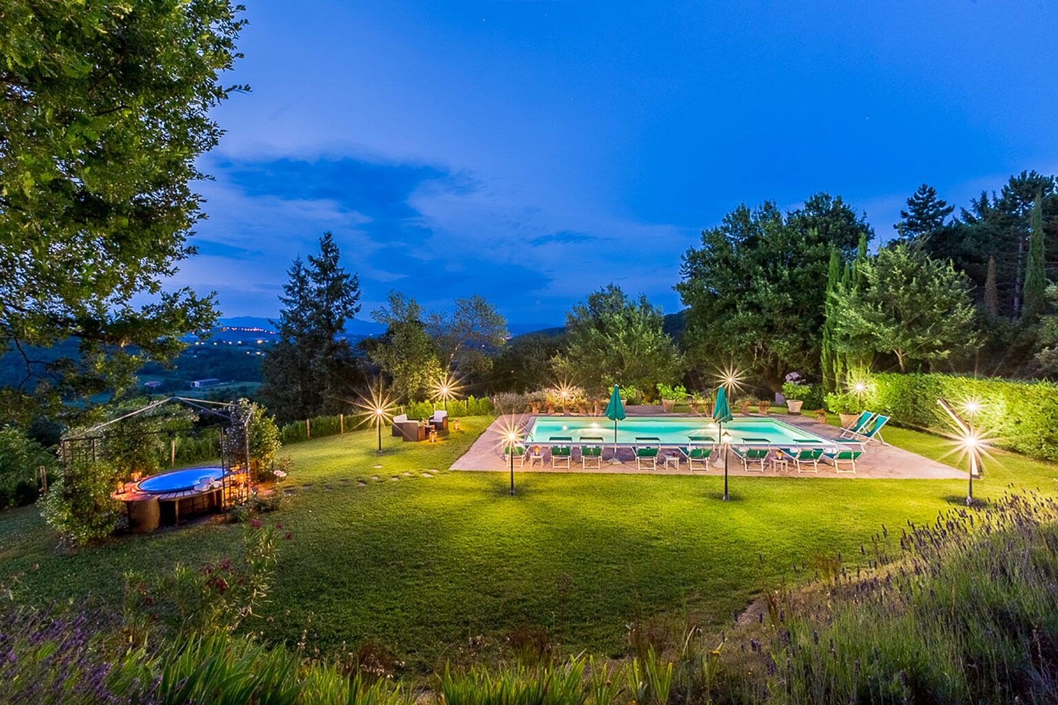 Scenic outdoor pool area at dusk, surrounded by lush greenery and illuminated by warm lights, featuring lounge chairs and a trampoline in a tranquil setting. | Only on SopranoVillas