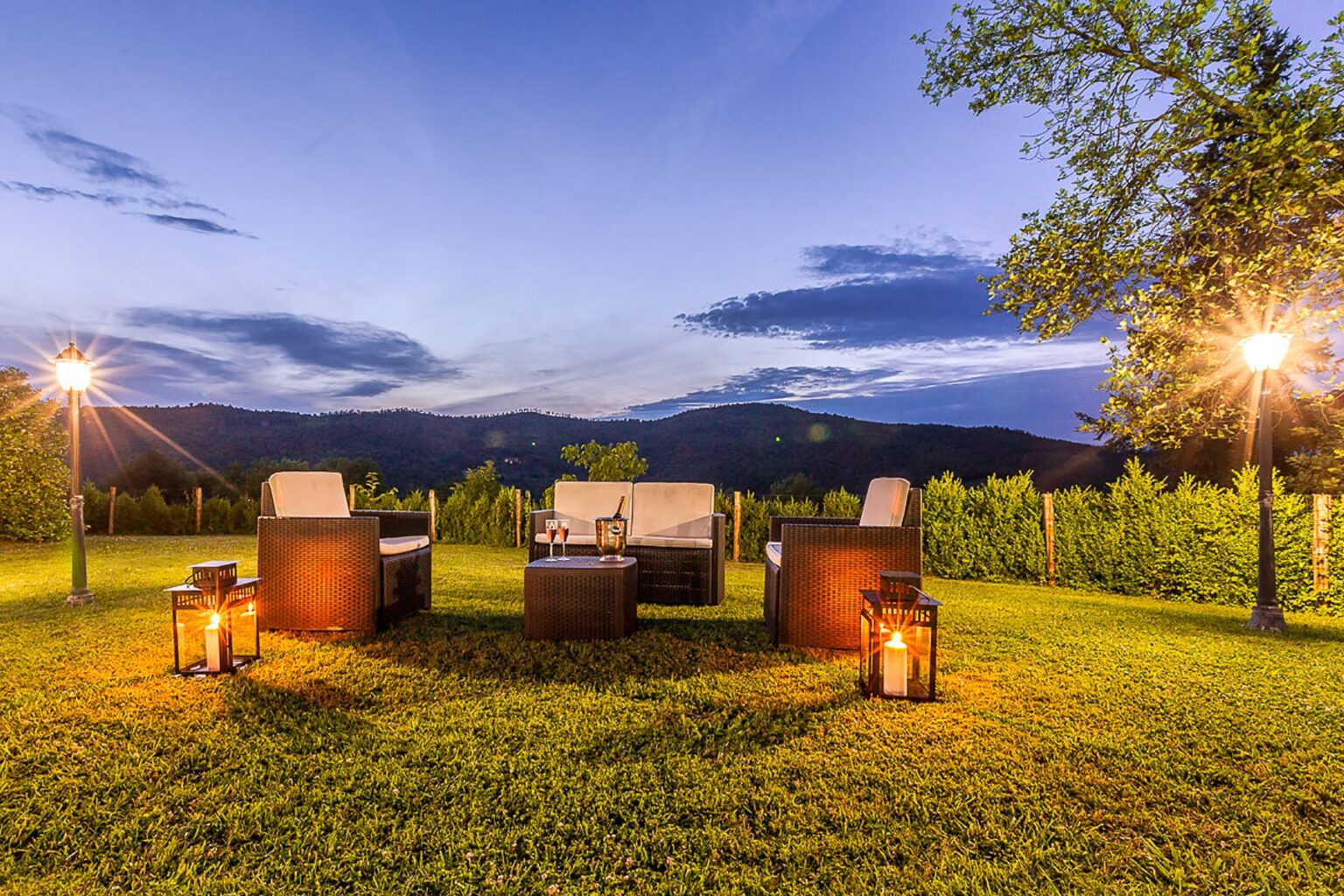 Cozy outdoor seating area with wicker chairs and a coffee table, illuminated by lanterns, set against a scenic mountain backdrop at dusk. | Only on SopranoVillas