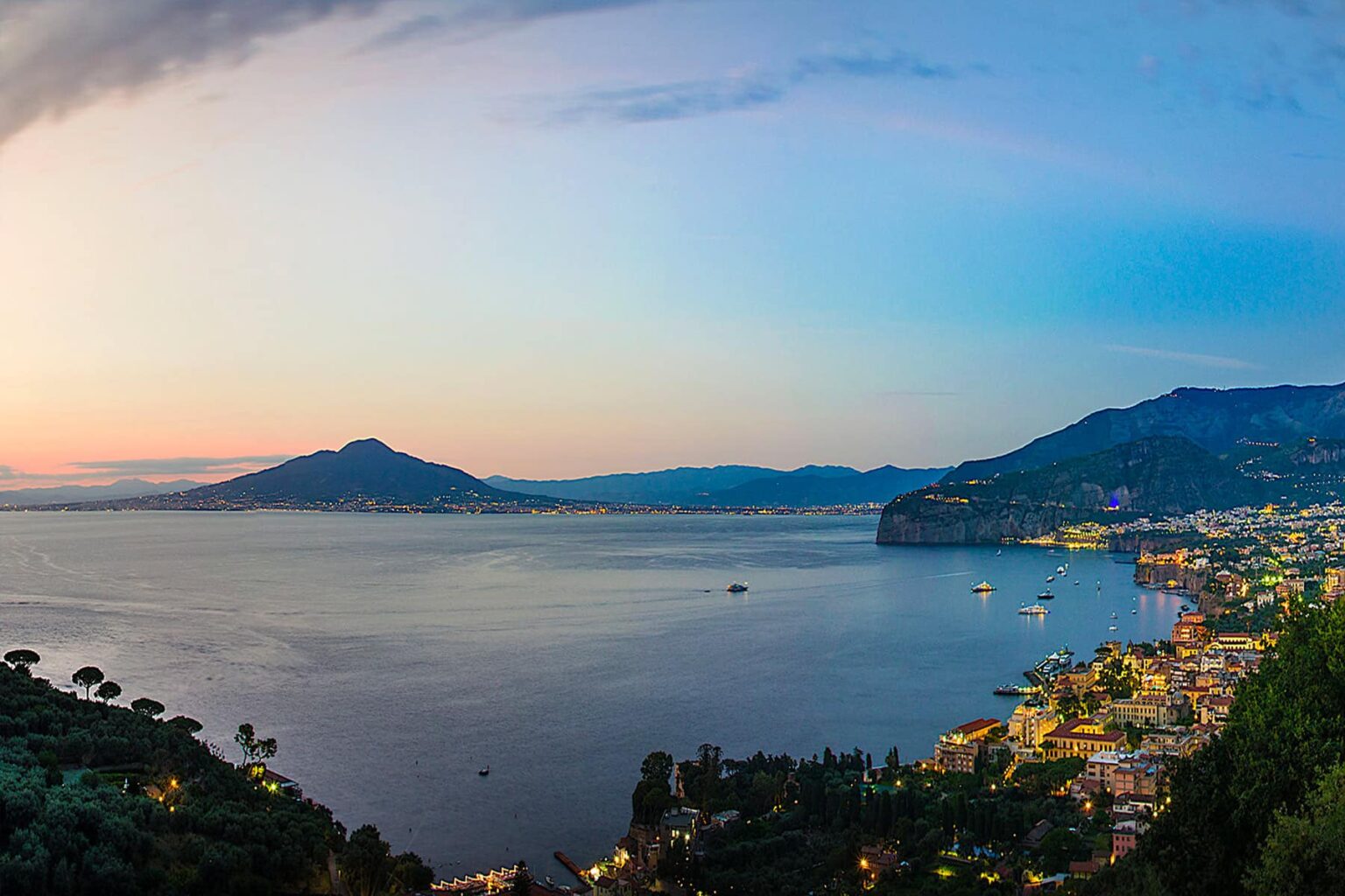 Panoramic view of the Bay of Naples at sunset, featuring Mount Vesuvius in the background and the coastal town of Sorrento with colorful buildings along the shoreline. The tranquil sea reflects the vibrant colors of the sky, creating a picturesque landscape. | Only on SopranoVillas