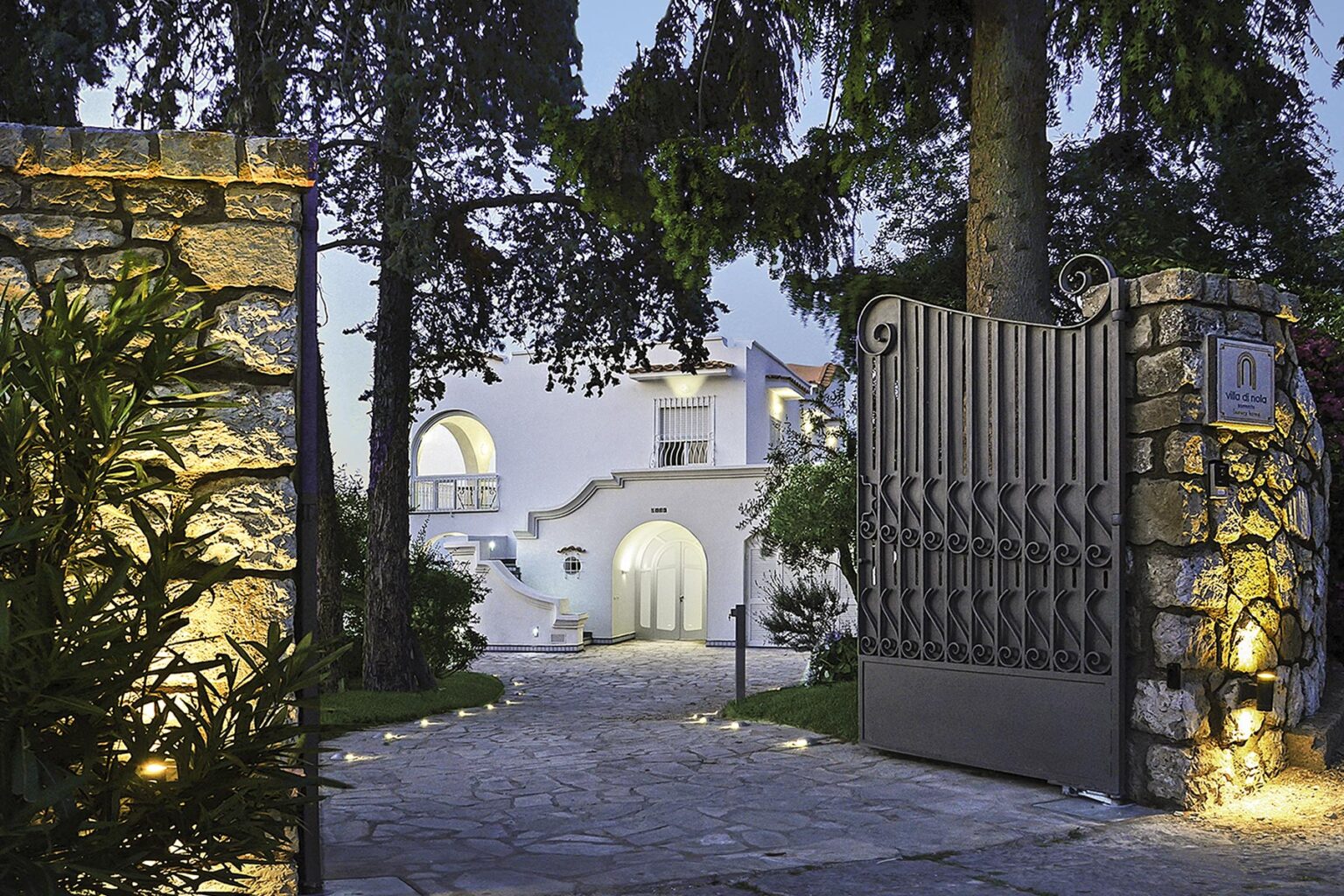 Elegant entrance of a modern villa, featuring a stone wall and decorative gate, surrounded by lush greenery and illuminated pathways leading to the main building. | Only on SopranoVillas