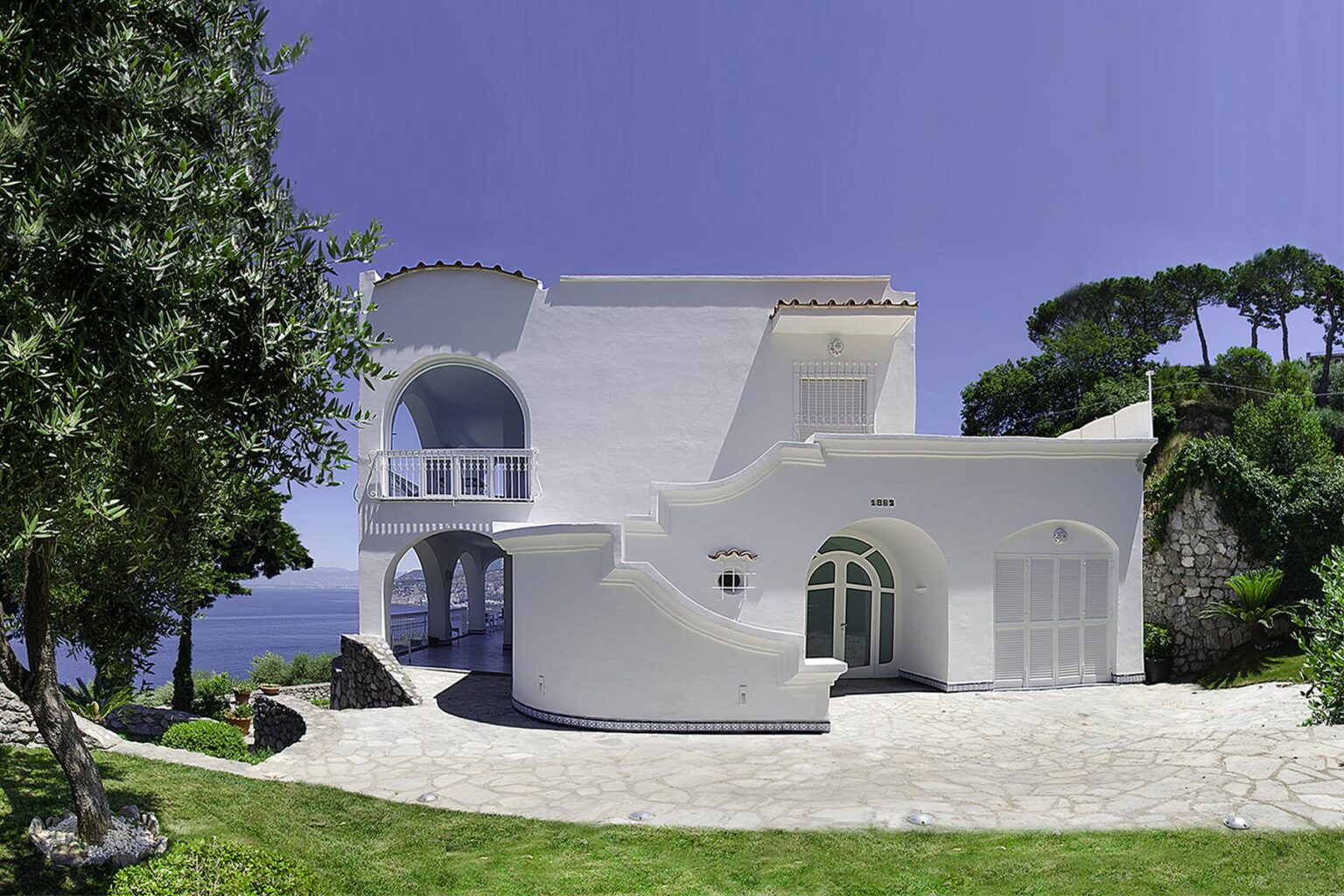 Modern white villa surrounded by lush greenery, featuring unique architectural design and panoramic views of the ocean under a clear blue sky. | Only on SopranoVillas
