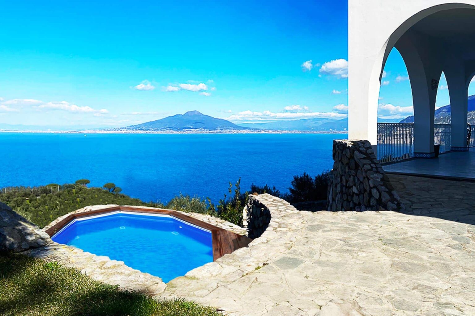 Scenic view of a luxury villa with a private pool overlooking the ocean and mountains, featuring clear blue skies and lush greenery. | Only on SopranoVillas