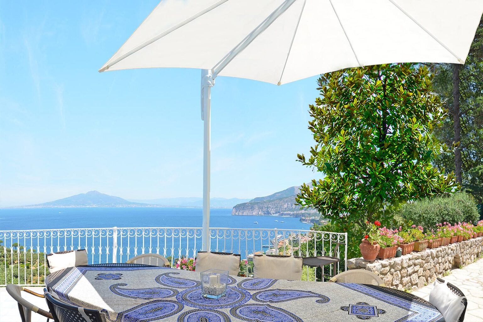 Outdoor dining area with a large white umbrella overlooking a scenic ocean view and lush greenery. The table features a decorative blue mosaic design, creating a relaxing atmosphere perfect for alfresco meals. | Only on SopranoVillas