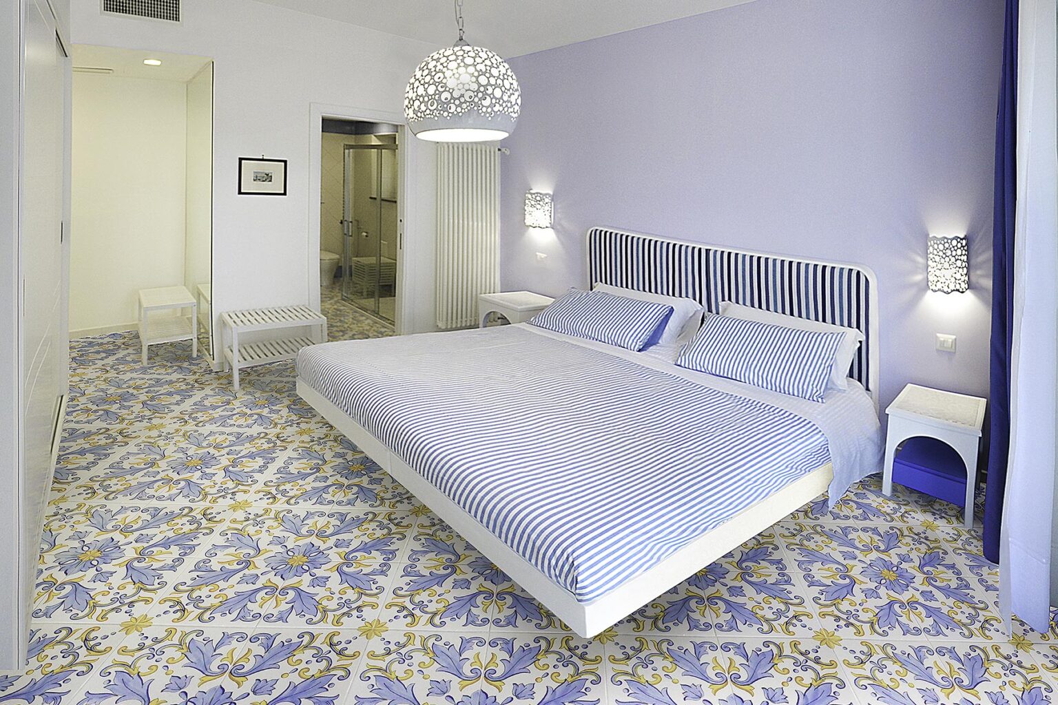 Modern bedroom with a large bed, featuring blue and white bedding, elegant lighting, and patterned tile flooring in shades of purple and yellow. The room has a serene atmosphere with minimalist decor and ample natural light. | Only on SopranoVillas