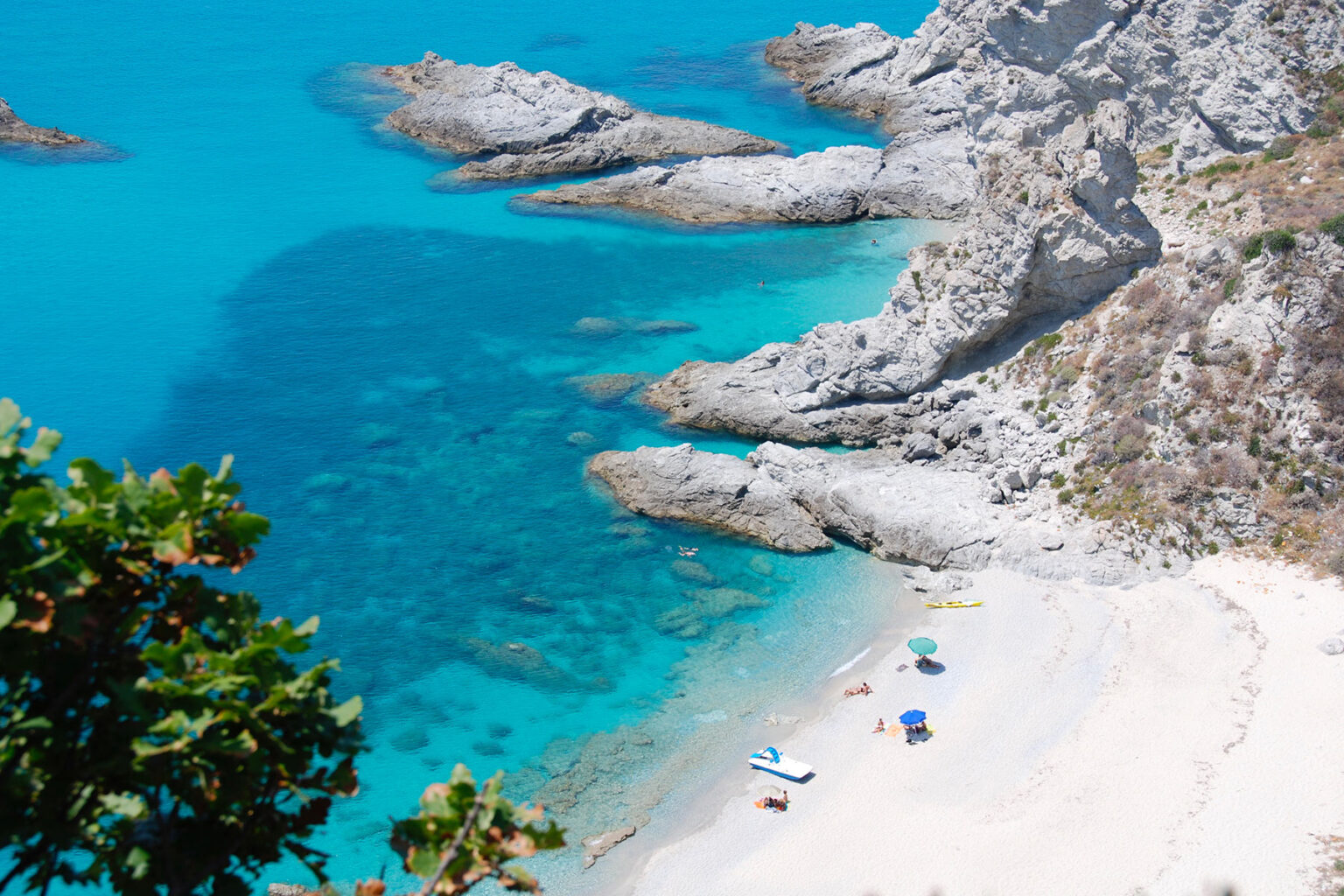 Aerial view of a serene beach with turquoise waters, rocky cliffs, and sunbathers enjoying the sun on colorful umbrellas and beach towels. | Only on SopranoVillas