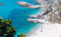 Aerial view of a serene beach with turquoise waters, rocky cliffs, and sunbathers enjoying the sun on colorful umbrellas and beach towels.