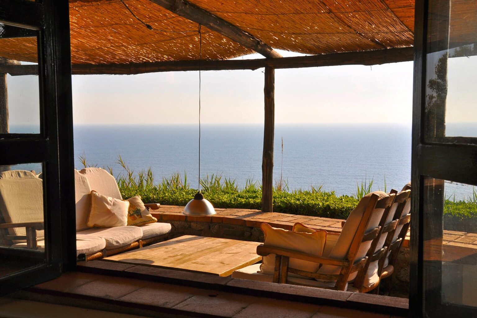 Cozy seating area with a wooden deck overlooking a tranquil ocean view, framed by a rustic structure and natural light. Ideal for relaxation and enjoying scenic coastal landscapes. | Only on SopranoVillas