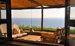 Cozy seating area with a wooden deck overlooking a tranquil ocean view, framed by a rustic structure and natural light. Ideal for relaxation and enjoying scenic coastal landscapes.