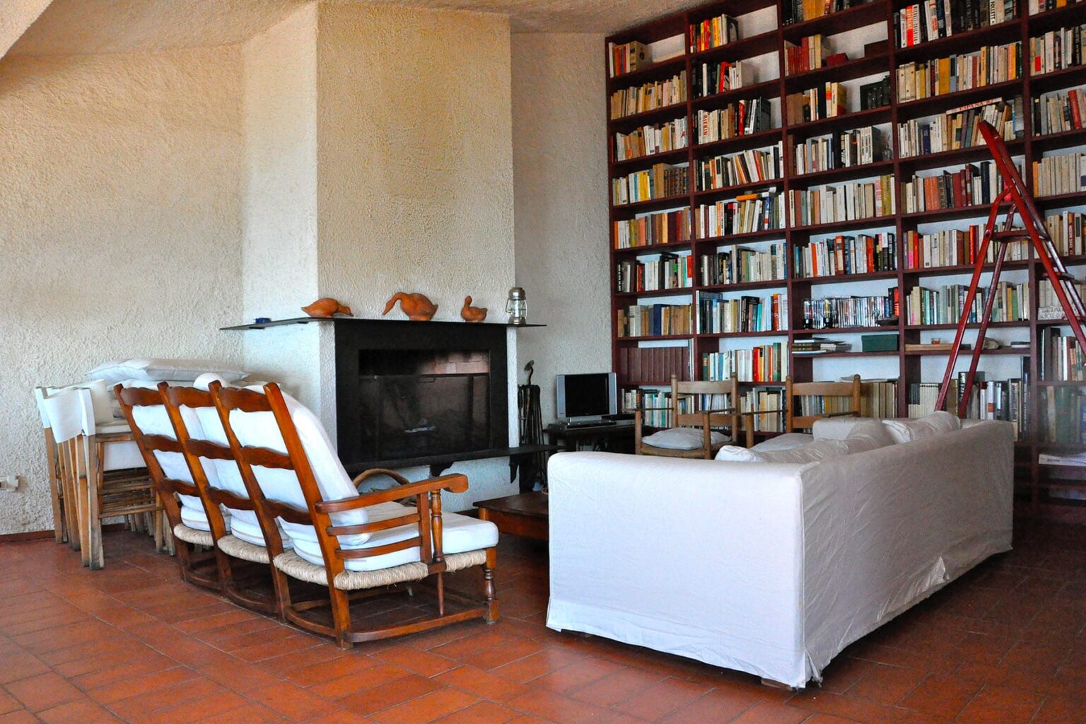Cozy living room featuring a large bookshelf filled with books, a modern fireplace, and comfortable seating arrangements including white and wooden chairs. The warm tile floor adds to the inviting atmosphere. | Only on SopranoVillas