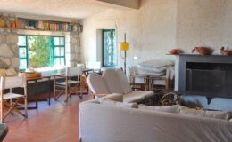 Cozy living room featuring a stone wall, large windows with natural light, comfortable seating, and a rustic wooden table, creating a warm and inviting atmosphere.