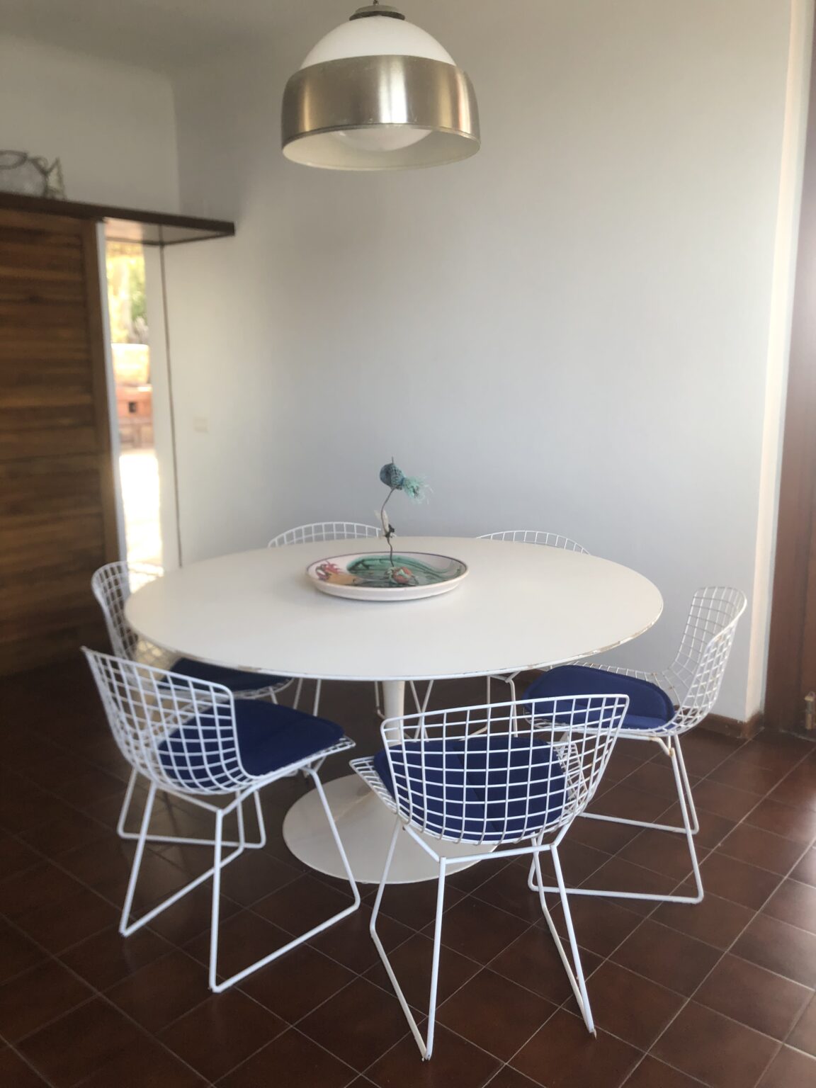 Modern dining area featuring a round white table surrounded by wire-frame chairs with blue cushions. The space includes a stylish pendant light and a decorative bowl on the table, set against a neutral wall and tile floor. | Only on SopranoVillas