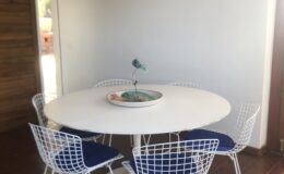 Modern dining area featuring a round white table surrounded by wire-frame chairs with blue cushions. The space includes a stylish pendant light and a decorative bowl on the table, set against a neutral wall and tile floor.