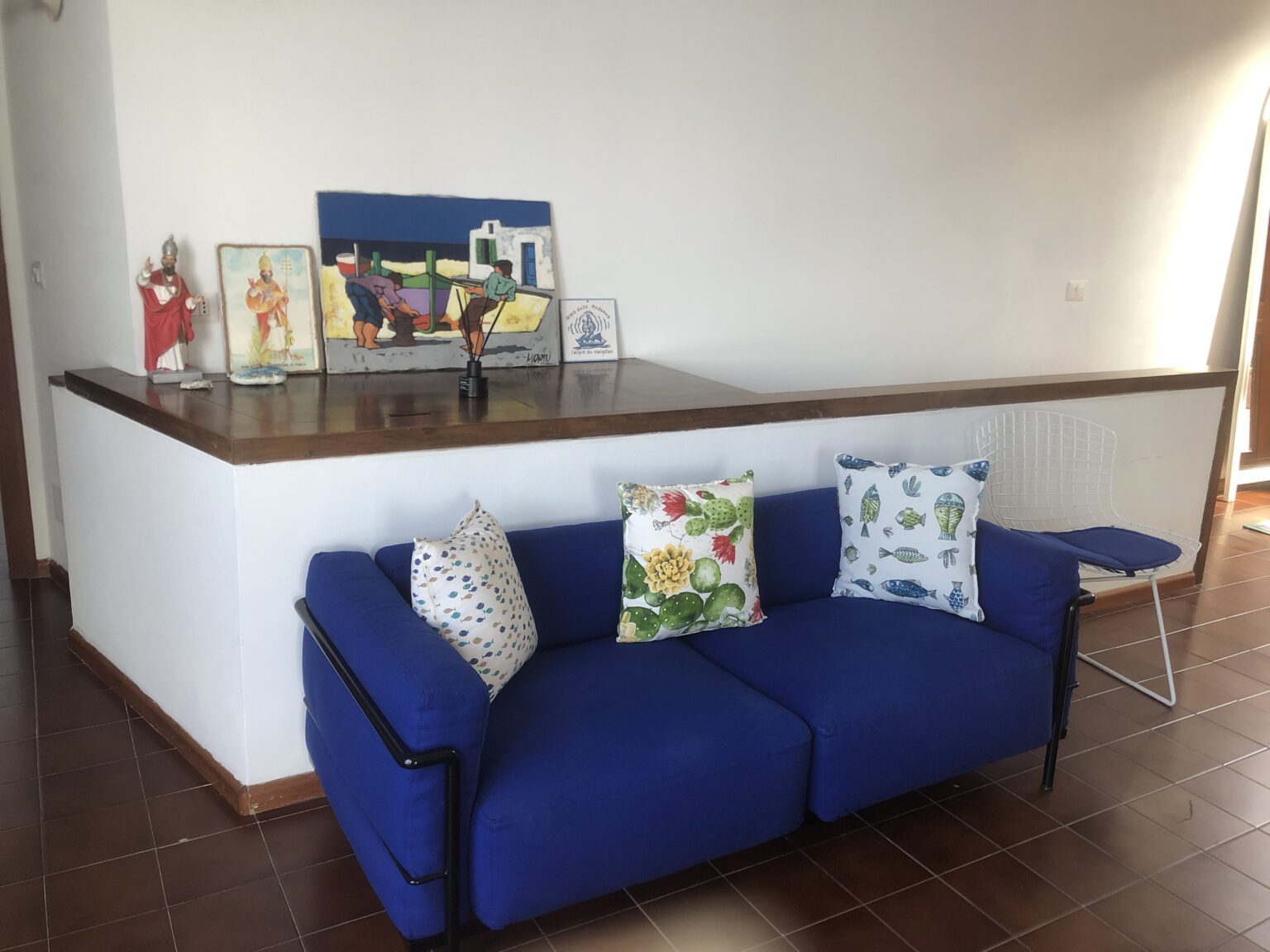 Bright living room featuring a modern blue sofa adorned with decorative pillows, a wooden bar counter, and vibrant artwork on the wall. A white wire chair complements the space, creating an inviting atmosphere. | Only on SopranoVillas