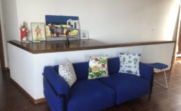 Bright living room featuring a modern blue sofa adorned with decorative pillows, a wooden bar counter, and vibrant artwork on the wall. A white wire chair complements the space, creating an inviting atmosphere.