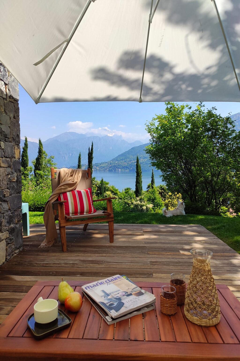 Cozy wooden chair with a red and white striped cushion, set on a wooden deck overlooking a serene lake and mountains, surrounded by lush greenery and trees. Ideal for relaxation in a picturesque outdoor setting. | Only on SopranoVillas
