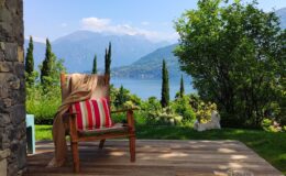 Cozy wooden chair with a red and white striped cushion, set on a wooden deck overlooking a serene lake and mountains, surrounded by lush greenery and trees. Ideal for relaxation in a picturesque outdoor setting.