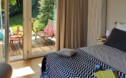 Cozy bedroom with a patterned black and white blanket, featuring a comfortable bed and large glass doors opening to a sunny outdoor patio with seating.